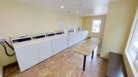 This shared laundry facility is a clean, efficiently organized space designed to accommodate high-volume laundry needs. The room is lined along one wall with an extensive row of white top-load washing machines and matching dryers, providing ample capacity for multiple users simultaneously. Each machine features a digital control panel, making operation straightforward and user-friendly. A spacious granite-topped folding table runs centrally through the room, offering a generous surface for sorting and folding laundry. The flooring throughout is durable, earth-toned ceramic tile that is easy to maintain and clean. Soft yellow-painted walls give the room a bright, welcoming atmosphere, complemented by recessed ceiling lighting that ensures the space is well-lit at all times. A window along the far wall allows natural light to filter in, adding to the room's open and airy feel. Ventilation is supported by ceiling-mounted HVAC registers, keeping the environment comfortable. The overall layout is practical and spacious, with clear circulation paths that allow multiple users to move through the room without congestion. This laundry facility is a functional and well-maintained amenity, thoughtfully designed for convenience and ease of use.