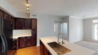 Located in Humble, TX, this single-floor residence offers a well-designed, functional layout that blends comfort and practicality throughout its living spaces. The open-concept kitchen serves as the heart of the home, showcasing rich dark wood cabinetry that extends to the ceiling for generous storage, light quartz countertops, and a large center island equipped with a deep single-basin stainless steel sink and a sleek industrial-style pull-down faucet. Track lighting illuminates the kitchen workspace, while black appliances—including a refrigerator, range, and microwave—add a cohesive, modern touch. Warm hardwood-style flooring runs through the kitchen and flows into the adjoining living area, which transitions to plush neutral carpeting. The living space provides an open, airy feel with a view directly to the front entry. The bedrooms are finished with soft carpet, crown molding, and ceiling fans, lending a refined yet cozy atmosphere. Natural light enters through horizontal-blind-fitted windows, offering a connection to the outdoors. The bathroom features a granite-topped vanity with dark wood cabinetry, a tub-shower combination, and direct access to a walk-in closet with built-in shelving and hanging rods—providing practical, organized storage. Neutral gray walls throughout create a clean, versatile backdrop.