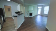 This single-story home showcases an open-concept design that seamlessly connects the kitchen and living areas into one cohesive, spacious layout. Dark hardwood flooring runs throughout, providing a rich contrast against the soft sage-green walls and crisp white trim and baseboards, creating a clean, welcoming ambiance.

The kitchen features a peninsula with a dark countertop, offering casual seating or prep space, along with white cabinetry above. This setup allows for easy interaction between the kitchen and the adjoining living area, making the space well-suited for entertaining.

The living area serves as the focal point of the home, anchored by a classic white-mantled fireplace with a tile surround. Above the mantle, a recessed panel adds architectural interest to the wall. Natural light fills the room through a glass-panel entry door with a transom window above, brightening the space and creating an inviting atmosphere.

Crown molding adds a refined touch throughout, while the open sightlines and airy flow between spaces make this home feel generous and functional. The overall aesthetic is polished yet approachable.