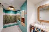 The Paws Spa is a thoughtfully designed pet grooming facility that combines bold aesthetics with highly functional features. The space is defined by its striking wall-to-wall tiling in a vivid checkerboard pattern of turquoise and amber, accented by a decorative band of patterned white tiles — creating a lively, resort-inspired atmosphere. The walk-in shower area is generously sized, equipped with dual brass hand-held shower fixtures and a built-in tiled bench, making pet bathing both convenient and comfortable. The shower floor features slip-resistant mosaic tile, while a floor drain ensures easy cleanup. A recessed niche built into the tiled accent wall provides storage for grooming supplies, accompanied by posted Paws Spa guidelines and occupancy information. Gold-tone leash hooks line the clean white wall opposite, adding both practicality and charm. The vanity area includes a marble-topped counter with an undermount sink, brass faucet fixtures, and richly patterned cabinetry in a bold geometric textile design, topped by a warm wood-framed mirror. A gold flower-shaped flush-mount ceiling fixture casts warm, ambient light throughout. The terrazzo-style flooring with a central floor drain completes this well-appointed, whimsical pet spa experience.
