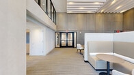 Located at Reston Row Plaza in Reston, Virginia, this sophisticated single-floor commercial space offers a generous, open layout designed to accommodate both collaborative and private work settings. The interior is defined by soaring double-height ceilings adorned with a striking slatted white ceiling feature, which lends a sense of architectural refinement and visual depth throughout the main lounge area.

Floor-to-ceiling glass walls and large-format windows flood the space with abundant natural light while framing expansive views of the surrounding Reston Row development. Neutral-toned carpet flooring anchors the space and complements a cohesive palette of warm beiges, whites, and natural wood tones.

The main lounge is furnished with high-backed booth-style seating, intimate round tables, and a curated mix of armchairs and sofas, creating a variety of seating configurations ideal for casual meetings or focused work. A quieter seating zone is set against textured wall paneling, offering a more enclosed and acoustically sheltered environment.

A dedicated private meeting room features a sleek rectangular conference table and upholstered chairs, framed by crisp white cabinetry and a full glass wall that opens to views of an outdoor terrace. Natural wood paneling near the entry area adds warmth and texture. Direct access to the outdoor terrace is provided via glass-paned doors, further enhancing the connection between the interior and the surrounding plaza environment.
