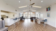 Located at 122 Jessup Rd in Henderson, Nevada, this single-floor professional office space serves as the leasing and management hub for The Pines community, operated by Fusion Property Management. The space is thoughtfully designed to balance functionality with a welcoming atmosphere.

The entrance greets visitors with an abundance of natural light streaming through tall, floor-to-ceiling windows and transom panes, offering views of the surrounding landscaping and pool area. The main open-plan area features soaring vaulted ceilings with multiple skylights that flood the interior with daylight, complemented by ceiling fans and recessed lighting. Rich wood-look flooring runs throughout, adding warmth to the bright white walls.

The main work area is equipped with sleek workstations and bar-height seating, ideal for client consultations. A comfortable lounge zone with upholstered seating provides a relaxed setting for guests. The kitchen area is fully appointed with white shaker cabinetry, a gray subway tile backsplash, stainless steel appliances including a refrigerator and range, and a white quartz peninsula with bar stools. Two bathrooms serve the space, one featuring accessible fixtures with grab bars and tile flooring, and the other a clean, modern design with a wide vanity. Colorful artwork adorns the walls throughout, contributing to a professional yet inviting office environment.