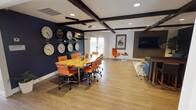 This thoughtfully designed single-floor space blends modern aesthetics with warm, rustic elements to create a versatile and inviting environment. Exposed dark wood ceiling beams run throughout, providing architectural character against bright white ceilings, while light hardwood flooring ties the various zones together seamlessly.

The centerpiece of the space is a generously sized conference area anchored by a striking live-edge wood table surrounded by vibrant orange rolling chairs. A bold navy accent wall serves as a dramatic backdrop, adorned with an eye-catching collection of world time zone clocks. Sleek pendant lighting overhead adds a contemporary touch, complementing the natural light that filters in through windows.

Adjacent to the conference area is a comfortable lounge zone featuring a large sectional sofa in a rich steel blue, set atop a plush shag rug. A wood-paneled feature wall hosts a large flat-screen television, making this area ideal for casual gatherings or media viewing. A metallic wall sculpture adds artistic flair to the navy-painted lounge wall.

A dedicated reception or workstation area is visible in the open-plan layout, equipped with desktop computers and a dark wood counter, providing a functional hub for administrative tasks. The walls throughout display a curated gallery of framed artwork, contributing to the space's creative and professional ambiance.