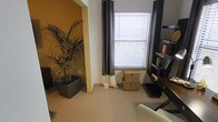 This single-floor space offers a warm and welcoming atmosphere, highlighted by a carefully curated interior design. The room visible features plush carpeted flooring in a neutral tone, contributing to a cozy and comfortable ambiance throughout. A striking arched alcove with a bold golden-yellow accent wall serves as a focal point, adding depth and character to the space. The alcove is complemented by a tall, lush tropical potted plant in a sleek dark planter, introducing a natural, organic element to the interior. Natural light filters generously through a large window dressed with elegant dark and cream-toned curtains paired with horizontal blinds, offering both style and privacy control. Woven storage baskets are neatly positioned beneath the window, blending practical storage with decorative charm. A dark wood bookshelf along one wall provides ample display and storage space, adding a refined, organized touch to the room. The overall design palette balances warm earth tones with neutral hues, creating an inviting and functional living environment. The space is well-appointed and thoughtfully arranged to maximize both comfort and aesthetic appeal.