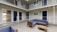 This courtyard serves as a central, open-air gathering space enclosed on multiple sides by a two-story building. The architecture features light neutral-toned vertical siding, white-painted exposed beam ceilings along the covered walkways, and crisp white grid-panel railings that line both the ground-level perimeter and the upper-floor balcony corridors. Bold navy blue entry doors provide a striking contrast against the pale façade and mark individual unit entrances accessible from both levels.

The courtyard floor is finished in smooth, light-toned concrete, providing a clean and durable surface throughout. Multiple seating groupings are distributed across the space, each consisting of teak-framed outdoor sofas, sectionals, and low coffee tables with blue-grey cushions, creating several distinct areas for gathering or relaxation. The open sky above allows abundant natural light to flood the courtyard throughout the day.

Ground-floor units are accessible directly from the courtyard, while upper-floor units are reached via stairways that connect to the second-level walkway. The wraparound balcony corridor on the upper floor overlooks the courtyard below, reinforcing the communal nature of the space. The overall ambiance is clean, modern, and inviting, with a consistent neutral and white color palette that lends a fresh, well-maintained character to the entire courtyard.
