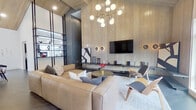 This single-floor communal space showcases a sophisticated contemporary design anchored by dramatic vaulted ceilings clad in warm wood planking, creating an immediately striking architectural impression. Large-format dark tile flooring flows seamlessly throughout, grounding the open layout and connecting each functional zone.

The lounge area is appointed with a generous tan leather sectional sofa, sculptural accent chairs, and a rounded coffee table set atop a plush area rug. A full-height wood-paneled feature wall hosts a wall-mounted television flanked by circular accent lighting and a low floating media console. Overhead, a multi-globe brass chandelier adds a refined mid-century modern touch.

Transitioning deeper into the space, a dedicated dining area centers around a long natural wood table with black-framed chairs beneath a substantial rectangular pendant light. An adjacent built-in media unit and a bar-height counter with seating extend the entertaining possibilities. Black-framed glass partitions define a private room nearby, adding functional versatility.

A bright entry foyer welcomes visitors with pool views through oversized windows and a sidelight door, while a separate poolside room features built-in navy cabinetry with a sleek countertop and expansive windows framing outdoor views. Natural light permeates throughout, reinforcing the space's open, airy ambiance.