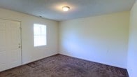 This single-story space includes a simply designed room finished with neutral, light-colored walls that create a clean and versatile backdrop. The flooring consists of wall-to-wall carpet in a medium brown tone, adding a degree of warmth and comfort underfoot. A window fitted with horizontal blinds allows natural light to filter into the room, brightening the interior while offering basic privacy control. The ceiling features a textured finish, commonly known as a popcorn or acoustic texture, and is outfitted with a centrally positioned flush-mount light fixture that provides ambient overhead lighting. White baseboards run along the perimeter of the room, providing a crisp contrast against the carpeted floor. A visible HVAC vent is integrated into the wall near the ceiling, indicating a ducted climate control system. The room's layout is open and unobstructed, offering flexibility for a variety of uses. The overall aesthetic is straightforward and functional, with a neutral palette throughout that would accommodate a range of furnishing styles and personal preferences.