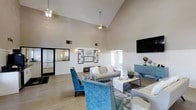 This single-floor clubhouse offers an inviting and well-appointed space designed to foster community and relaxation. The main lounge area immediately impresses with its dramatic vaulted ceilings, which lend a sense of grandeur and openness to the interior. Light-toned walls, accented by classic white wainscoting, create a clean and polished aesthetic throughout.

The lounge is furnished with a comfortable neutral sofa and complemented by bold teal accent chairs, adding a pop of color against the light hardwood-style flooring. A patterned area rug anchors the seating arrangement around a sleek, modern coffee table, while a wall-mounted flat-screen television serves as a central entertainment focal point. Pendant lighting fixtures hang from the high ceiling, providing warm, ambient illumination alongside natural light filtering in through the windows.

A teal sideboard along the wall offers additional storage and display space, decorated with tasteful accessories. Framed artwork and a decorative pin board add personality to the walls, contributing to a community-oriented atmosphere. An adjacent mirrored area is visible to one side, suggesting additional functional space nearby. Overall, this clubhouse combines thoughtful design with practical amenities, making it an ideal gathering spot for residents and guests alike.