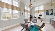 Located at 100 Dalton Ln in Charlottesville, VA, this single-room living space offers a thoughtfully designed interior that balances comfort, functionality, and refined aesthetics. The room is defined by impressively tall ceilings adorned with crisp crown molding, which amplify the sense of openness and grandeur. Large, multi-pane windows line the walls, dressed with neutral-toned striped valances, allowing an abundance of natural light to flood the space while offering views of the surrounding landscape and outdoor railings. The light-toned tile flooring adds a clean, airy quality underfoot. A striking cluster pendant chandelier with amber glass globes serves as a focal point, casting a warm, inviting glow. The seating area centers around a grouping of mid-century modern upholstered chairs arranged around a round white coffee table atop a vibrant teal area rug, creating an engaging conversational setting. A built-in workstation along one wall features rich wood cabinetry with granite countertops, a glossy white tile backsplash, and pendant lighting — ideal for a beverage or hospitality station. A standing desk with computer monitors along the windowed wall provides a dedicated workspace. Contemporary artwork in cool blue tones completes the sophisticated, welcoming atmosphere of this well-appointed space.
