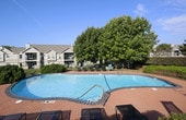 This single-story amenity facility offers residents a well-rounded outdoor and indoor retreat. At the heart of the space is a generously sized swimming pool with crystal-clear blue water, framed by a brick-paved deck lined with lounge chairs and Adirondack seating. Mature trees provide natural shade along the pool perimeter, complemented by manicured landscaping and a secure fencing system. A multi-building residential backdrop is visible from the pool area, reinforcing the community setting.

The facility includes an architecturally distinctive glass-enclosed sunroom, featuring a curved, arched roof with dark metal framing and expansive floor-to-ceiling windows that flood the interior with natural light. The sunroom is finished with rustic wood plank flooring and is furnished with wrought iron seating, creating an inviting indoor-outdoor atmosphere with direct views of the pool deck and surrounding greenery.

Access to the facility is provided through glass-paneled entry doors, with a tiled transitional entryway connecting the sunroom to adjacent interior rooms. Interior corridors feature tile flooring with decorative inlay patterns and standard white-paneled doors leading to utility and support spaces. The overall design balances functional amenity use with a comfortable, community-oriented ambiance.