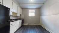 This single-floor unit is situated in a park side location, offering a well-organized layout across multiple rooms. The interior is unified by dark hardwood-style flooring that runs throughout, complemented by neutral gray and taupe wall tones and crisp white trim and baseboards. The living area features a tray ceiling with exposed beams, a large window that invites natural light, and a baseboard heating system. Each bedroom is generously proportioned, with multiple windows offering views of the surrounding neighborhood and ample wall space. The kitchen is functionally designed, equipped with white cabinetry, dark granite countertops, a tiled backsplash, a stainless steel sink, and black appliances including a refrigerator and gas range. A window above the kitchen area provides additional light and ventilation. The bathroom retains a classic aesthetic, showcasing a black-and-white mosaic tile floor, white subway-tiled walls, a combination tub and shower, and a frosted window for privacy. Connecting hallways and doorways with white panel doors create a cohesive flow throughout the unit. Overall, the space presents a clean, updated interior with functional details suited for comfortable everyday living.