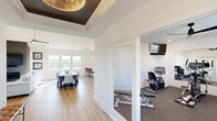 Located at 8495 Hoke Rd in Clayton, Ohio, this single-story community space is designed with a clean, contemporary aesthetic that prioritizes openness, natural light, and functional gathering areas. The entryway immediately establishes a refined tone with a dramatic tray ceiling accented by a bold dark inset and a statement gold chandelier, paired with warm luxury vinyl plank flooring that flows throughout the main areas.

The central great room is anchored by a striking dark accent wall housing a wall-mounted fireplace and flat-screen television. Soaring vaulted ceilings with a ceiling fan enhance the sense of spaciousness, while an abundance of windows along the perimeter frame sweeping outdoor views of the surrounding landscape, flooding the interior with natural light. A generous dining area with seating for a large group sits adjacent, complemented by counter-height bar seating along a side wall.

A well-equipped kitchenette features gray shaker-style cabinetry, crisp white countertops, a beverage refrigerator, microwave, and a coffee station, making it ideal for hosting gatherings or events. A dedicated fitness room with carpeted flooring, exercise equipment, and a wall-mounted monitor offers a convenient wellness amenity. A comfortable lounge alcove near the exterior door provides a welcoming seating area with curated artwork and direct access to an outdoor patio.