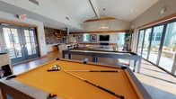 This thoughtfully designed single-floor amenity space blends modern style with warm, inviting materials to create a versatile gathering destination. The open layout flows seamlessly between distinct activity zones, all unified by rich wood-toned flooring, warm timber beam accents, and a neutral palette that gives the space a polished yet comfortable feel.

At the heart of the room, a full-size billiards table and a shuffleboard table take center stage, offering prime entertainment options. Adjacent to the game area, a well-appointed kitchen features a large central island with seating, a herringbone-patterned tile backsplash, floating wood shelves, stainless steel appliances, and bold pendant lighting — ideal for hosting gatherings or catered events.

The lounge area anchors around a dramatic floor-to-ceiling stacked stone fireplace with a wall-mounted television above, framed by soaring vaulted ceilings and an eye-catching sputnik-style chandelier. Comfortable mustard-toned seating and a substantial wood coffee table complete the relaxed atmosphere.

Floor-to-ceiling sliding glass doors flood the space with natural light and open directly onto an outdoor patio, seamlessly connecting interior and exterior living. Curated artwork, sculptural décor accents, and lush indoor plants add layers of visual interest throughout, making this an exceptionally well-appointed communal space.