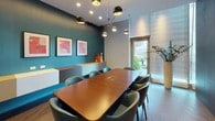 This conference room is a thoughtfully designed meeting space that balances professional functionality with contemporary style. At its center sits a substantial warm-toned wood conference table surrounded by comfortable teal upholstered chairs, providing ample seating for a full group. The room's design draws the eye with a bold teal textured accent wall, complemented by crisp white walls and rich dark hardwood flooring throughout.

Lighting is a standout feature, with a sculptural multi-arm pendant fixture fitted with globe-shaped spotlights casting a warm, focused glow over the table. Recessed ceiling lights provide supplemental illumination, ensuring the space is well-lit for productive meetings.

A wall-mounted flat-screen display is positioned for easy viewing from all seats, making the room well-suited for presentations and video conferencing. A floating credenza along the teal accent wall offers additional surface space, accessorized with decorative objects and framed artwork that add personality to the room.

One wall features a dramatic floor-to-ceiling dark panel with sleek brass handles, lending a sophisticated, refined character. Floor-to-ceiling windows fitted with horizontal blinds flood the space with natural light and offer views of lush outdoor greenery. A glass door provides direct access to an exterior courtyard, further enhancing the room's connection to the outdoors and creating an open, inviting atmosphere.