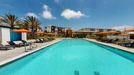 Located in San Pedro, CA, this single-level outdoor amenity space is designed to deliver a resort-like experience under the Southern California sun. At its centerpiece is a generously sized swimming pool with crystal-clear turquoise water, framed by a wide concrete sun deck lined with sleek charcoal lounge chairs and vibrant orange market umbrellas and shade sails that add bold pops of color throughout the space.

Adjacent to the pool, a covered outdoor lounge pavilion provides a comfortable retreat from the sun. The structure features a corrugated metal canopy supported by steel columns, with seating areas and fire pit-style tables that encourage gathering and relaxation. Floor-to-ceiling glass walls connect the interior amenity building — clad in warm wood paneling — to the outdoor environment, creating a seamless indoor-outdoor flow.

The surrounding landscape is thoughtfully designed with drought-tolerant native plantings, including ornamental grasses, agaves, succulents, and flowering purple sage, reflecting a commitment to sustainable design suited to the Southern California climate. Tall palm trees and established hedgerows provide natural privacy screening. The overall ambiance is modern, open, and inviting, ideal for outdoor recreation and leisurely enjoyment year-round.