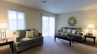 This single-floor residence centers around a welcoming living room finished with wall-to-wall carpeting in a neutral tone, creating a warm and cohesive atmosphere. The room is bathed in natural light, courtesy of a large sliding glass door fitted with vertical blinds that lead to an outdoor area, complemented by an additional window along the adjacent wall. The light-colored walls and textured ceiling contribute to an open, airy feel throughout the space. The layout comfortably accommodates multiple seating arrangements, making it well-suited for both relaxed living and casual entertaining. A decorative sunburst wall accent adds a stylistic focal point to the room, enhancing the overall aesthetic. The ceiling-mounted HVAC vent indicates a central air and heating system, ensuring year-round comfort. The neutral color palette throughout provides a versatile backdrop, allowing for a variety of interior design styles. Overall, the living area strikes a balance between functionality and comfort, with generous natural light and practical finishes defining the character of the space.