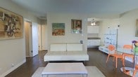This two-floor space presents a clean, minimalist aesthetic defined by a neutral color palette of soft whites and warm beiges. The main living area features dark hardwood flooring that contrasts effectively with the light-toned walls and furnishings. A contemporary white tufted sofa serves as the focal point of the living space, complemented by a sleek white coffee table positioned atop a light area rug. Decorative artwork adorns the walls, adding visual interest to the otherwise understated design. The living area flows openly into the kitchen, which is equipped with white cabinetry, open shelving, and a countertop that provides ample workspace. A ceiling fan with a light fixture is installed in the kitchen area, promoting airflow and functionality. White shelving units offer additional storage and display space. The textured ceiling adds subtle dimension throughout. Hallway access visible from the main living space suggests additional rooms beyond, while the open-plan layout between the living and kitchen zones creates a cohesive and practical environment. The overall ambiance is bright, airy, and simply styled.
