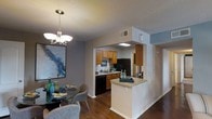 This single-floor residence offers a thoughtfully designed open-concept layout that seamlessly connects the kitchen and dining areas, creating a cohesive and inviting living environment. Rich hardwood flooring flows throughout, adding warmth and continuity to the space.

The kitchen is well-appointed with honey-toned wood cabinetry, dark-finish appliances including a refrigerator and dishwasher, and a stainless steel sink set beneath a functional countertop workspace. A peninsula counter extends from the kitchen, providing additional prep space and serving as a natural transition point between the kitchen and the rest of the living area. Recessed lighting above the kitchen ensures the space is well-lit and practical.

Adjacent to the kitchen, the dining area is anchored by a round glass-top table paired with contemporary upholstered chairs, creating an intimate setting for meals. A stylish multi-bulb chandelier hangs overhead, adding a touch of elegance to the dining nook. A large abstract wall art piece in cool blue tones brings a splash of color and personality to the space.

The textured ceiling and neutral wall tones throughout provide a clean, versatile backdrop that complements the warm flooring and cabinetry. Overall, the space strikes a balance between functionality and comfort, making it a welcoming everyday living environment.