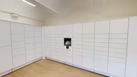 This single-floor space is purpose-built as a package locker room, designed to provide a clean, efficient, and organized environment for parcel management. The room is lined on multiple walls with floor-to-ceiling white smart locker units, offering an extensive array of compartments in varying sizes to accommodate packages of different dimensions. A centrally positioned touchscreen interface panel is embedded within the locker bank, enabling users to access their deliveries conveniently and securely.

The flooring throughout the space consists of light wood-tone planks, lending a warm contrast to the crisp white cabinetry. The ceiling is finished in a neutral tone and features recessed linear lighting that evenly illuminates the room. A vaulted ceiling detail adds a subtle architectural interest overhead.

A glass-paneled entry door with a sidelight allows natural light to filter into the space, offering a partial view of an outdoor greenery area beyond. The overall design is minimal and functional, with clean lines and a neutral color palette that keeps the focus on accessibility and ease of use. The space is climate-controlled, with a wall-mounted HVAC vent visible near the locker units.
