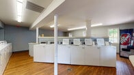 This laundry center is a single-floor facility designed to provide a functional and comfortable experience. The space is anchored by an extensive lineup of commercial-grade top-loading washers and dryers arranged along the walls, offering ample capacity to accommodate high-volume use. A dedicated utility sink station provides added convenience for pre-treating or hand-washing garments. The flooring throughout the facility consists of warm hardwood, lending a welcoming character to what is typically a purely utilitarian space. Structural support columns are integrated throughout the open floor plan, preserving a sense of spaciousness. A service counter near the entrance serves as an informational or assistance station. The waiting area is thoughtfully furnished with cushioned lounge chairs and ottomans, accompanied by a small side table, allowing users to relax comfortably. Wire laundry carts on wheels and a freestanding garment rack are available for sorting and organizing clothes. Multiple windows along the walls invite natural light and provide views of the surrounding greenery outdoors. Overhead fluorescent lighting ensures the entire space is consistently and evenly illuminated. A vending machine near the entrance offers added convenience. The interior walls are painted in a calm, muted blue-gray tone, contributing to a clean and orderly ambiance throughout the facility.