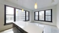 This contemporary single-floor space showcases a clean, open-plan design defined by a crisp white color palette and minimalist aesthetic. The interior is flooded with natural light through expansive black-framed windows that frame striking views of the surrounding urban skyline, creating a dynamic connection between the interior and the city beyond. Pendant lighting fixtures hang from the bright white ceiling, adding warm, focused illumination that complements the natural light throughout the day. A prominent island or countertop finished in polished white provides a generous work or gathering surface, flanked by sleek dark cabinetry with brushed metal hardware — a bold contrast that anchors the space with a modern, sophisticated feel. The open floor plan creates a seamless flow and a strong sense of spaciousness, making the interior feel airy and versatile. The neutral tones of the walls and flooring enhance the brightness of the space while maintaining a professional and refined ambiance. Overall, this space combines thoughtful design details with an abundance of natural light and commanding city views to deliver a polished, functional, and visually striking environment well-suited for a variety of uses.