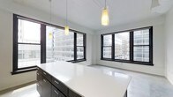 This contemporary single-floor space showcases a clean, open-plan design defined by a crisp white color palette and minimalist aesthetic. The interior is flooded with natural light through expansive black-framed windows that frame striking views of the surrounding urban skyline, creating a dynamic connection between the interior and the city beyond. Pendant lighting fixtures hang from the bright white ceiling, adding warm, focused illumination that complements the natural light throughout the day. A prominent island or countertop finished in polished white provides a generous work or gathering surface, flanked by sleek dark cabinetry with brushed metal hardware — a bold contrast that anchors the space with a modern, sophisticated feel. The open floor plan creates a seamless flow and a strong sense of spaciousness, making the interior feel airy and versatile. The neutral tones of the walls and flooring enhance the brightness of the space while maintaining a professional and refined ambiance. Overall, this space combines thoughtful design details with an abundance of natural light and commanding city views to deliver a polished, functional, and visually striking environment well-suited for a variety of uses.