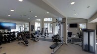 This two-floor fitness facility is thoughtfully designed to support a full range of workout routines. The main workout area is bright and open, featuring soaring ceilings and large windows that allow abundant natural light to fill the space, creating an energizing atmosphere. Recessed lighting throughout supplements the natural light, ensuring excellent visibility at any time of day.

The strength training zone is comprehensively equipped, including a multi-function cable machine, a variety of adjustable weight benches, and a full dumbbell rack offering a wide selection of free weights. Medicine balls and additional accessories are neatly organized and readily accessible. Wall-mounted mirrors reflect the open layout, enhancing the sense of spaciousness while allowing users to monitor their form during exercise.

The flooring transitions thoughtfully between zones, with durable, cushioned surfaces in key workout areas providing both safety and comfort. A wall-mounted flat-screen television adds to the overall experience. The neutral color palette of warm whites and soft grays throughout lends the facility a clean, modern aesthetic. The facility's layout is open and uncluttered, promoting ease of movement between equipment and creating a welcoming environment for users of all fitness levels.
