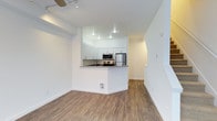 This two-floor residence showcases a clean, contemporary design characterized by bright white walls, wood-look flooring, and an efficient open-concept layout. The main living area flows seamlessly into a peninsula-style kitchen, which serves as the visual centerpiece of the space. The kitchen is equipped with stainless steel appliances, including a refrigerator and microwave, complemented by white shaker-style cabinetry and a dark countertop that provides a subtle contrast to the otherwise light palette. The peninsula doubles as a casual dining or prep area, enhancing both functionality and social interaction within the space. Recessed lighting in the kitchen ensures ample illumination, while the living area benefits from a ceiling fan with integrated track lighting, offering flexible light control. An arched passageway visible toward the rear of the main floor adds an architectural detail that softens the otherwise modern aesthetic and leads to additional areas of the home, including access to the second floor. The consistent flooring and neutral tones throughout create a cohesive, move-in-ready environment suited to a variety of interior styles.