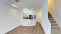 This two-floor residence showcases a clean, contemporary design characterized by bright white walls, wood-look flooring, and an efficient open-concept layout. The main living area flows seamlessly into a peninsula-style kitchen, which serves as the visual centerpiece of the space. The kitchen is equipped with stainless steel appliances, including a refrigerator and microwave, complemented by white shaker-style cabinetry and a dark countertop that provides a subtle contrast to the otherwise light palette. The peninsula doubles as a casual dining or prep area, enhancing both functionality and social interaction within the space. Recessed lighting in the kitchen ensures ample illumination, while the living area benefits from a ceiling fan with integrated track lighting, offering flexible light control. An arched passageway visible toward the rear of the main floor adds an architectural detail that softens the otherwise modern aesthetic and leads to additional areas of the home, including access to the second floor. The consistent flooring and neutral tones throughout create a cohesive, move-in-ready environment suited to a variety of interior styles.