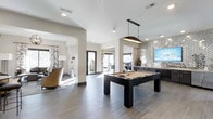 This contemporary amenity lounge is designed for both relaxation and recreation, offering a well-appointed, open-concept layout across a single floor. At the heart of the space sits a sleek, dark-framed billiards table, illuminated by a stylish pendant light that anchors the room with a modern aesthetic. Large-format tile flooring in a cool gray tone extends throughout, lending the space a clean, polished look.

The entertainment area features a striking herringbone-patterned accent wall in a metallic finish, serving as a sophisticated backdrop for a wall-mounted flat-screen display and a dark cabinetry media console below. Adjacent to this, an open archway leads into a comfortable lounge area furnished with plush seating, a floor lamp, and decorative drapery panels, creating a more intimate retreat within the broader space.

Natural light pours in through floor-to-ceiling glass doors at the far end, which open to an outdoor area, further enhancing the bright and airy atmosphere. The high white ceilings and neutral wall tones amplify the sense of spaciousness throughout. This thoughtfully designed space combines functionality and modern style, making it an ideal setting for socializing and leisure.