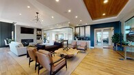 Located at 3040 Central Avenue in Billings, Montana, this single-story community amenity space combines contemporary design with a welcoming, upscale atmosphere. The main lounge area serves as the heart of the facility, featuring warm wood-look flooring, vaulted and tray ceilings with recessed lighting, and a striking wood-paneled ceiling accent. A dramatic floor-to-ceiling glass partition elegantly separates the lounge from the billiards room, which is bathed in natural light from multiple windows and anchored by a solid wood pool table. The open lounge is furnished with a mix of neutral sofas, mid-century modern accent chairs, and a live-edge wood coffee table, all set against a bold navy blue accent wall with decorative metallic panels. A compact kitchenette offers dark granite countertops, crisp white cabinetry, a mosaic glass tile backsplash in teal and silver tones, and modern appliances. The dedicated fitness center is well-appointed with cardio machines including treadmills and a stair climber, a multi-station cable machine, free weights, medicine balls, wall-mounted TVs, and large windows that bring in ample natural light. A shared laundry room with stacked and front-load machines completes the space, providing practical convenience for residents.