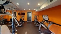 This single-floor fitness center is designed to provide a functional and energizing workout environment. The space is immediately distinguished by its bold orange walls, which lend a vibrant, motivating atmosphere throughout. Recessed ceiling lighting illuminates the room evenly, complemented by a textured white ceiling overhead.

The fitness center is divided into two connected areas accessible through a glass-paned interior door. The first area is dedicated to cardio equipment, including a stationary bike, an elliptical trainer, and a stair climber, all arranged along the walls to maximize floor space. A wall-mounted flat-screen television is positioned for easy viewing during workouts. Full-length mirrors on one wall enhance the sense of space and allow for form monitoring.

The second area houses additional cardio machines, including a treadmill, alongside a multi-station strength training machine and a free weight bench with dumbbells. This section features a glass exit door that opens directly to an outdoor green space, allowing natural light to fill the room and providing views of the surrounding landscape.

Durable rubber flooring covers both areas, offering safety and comfort underfoot. A water cooler and additional safety features, including clearly marked exit signage, are also present. The overall layout is practical and efficiently organized to support a full range of workout routines.
