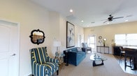 Academy Park Apartments presents a thoughtfully designed single-floor community space that combines comfort and style in an inviting setting. The main lounge area is appointed with an eye-catching mix of furnishings, including a plush tufted blue sofa, bold blue-and-yellow striped accent chairs, and a sleek glass coffee table, creating a cohesive and welcoming aesthetic. Recessed lighting throughout the ceiling ensures consistent, well-distributed illumination, while a ceiling fan adds functional comfort to the space. The walls, finished in a clean neutral tone, serve as an elegant backdrop for carefully selected décor, including a large framed abstract artwork and a distinctive quatrefoil-shaped decorative mirror. Natural light filters in through a window near the entrance, brightening the space and giving it an open, airy feel. A console table near the entryway, adorned with decorative accents and a mirror, adds a refined finishing touch. The carpeted flooring contributes warmth underfoot, complementing the overall inviting ambiance. This community lounge at Academy Park Apartments is designed to offer residents a comfortable and attractive space for relaxation and socializing.