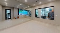 This single-level fitness studio is thoughtfully designed to support a wide range of exercise and wellness activities. The space features a clean, contemporary aesthetic defined by neutral-toned walls, crisp white baseboards, and light-colored flooring that lends a bright, open feel underfoot.

A defining architectural feature is the expansive floor-to-ceiling mirror wall, which visually amplifies the room and provides essential feedback for movement-based training, yoga, or dance. Recessed ceiling lighting is evenly distributed throughout, ensuring the space is consistently and effectively illuminated without harsh shadows.

A large wall-mounted display screen serves as a central focal point, enabling on-demand workout classes, virtual training sessions, or ambient visuals to enhance the exercise experience. Windows with black-framed trim allow natural light to filter in while offering views to adjacent areas, connecting the studio to its surroundings.

A dedicated nook near the entry houses fitness accessories including foam rollers, a BOSU balance trainer, and yoga blocks, keeping essential equipment organized and within easy reach. The bold black-framed entry door adds a striking contrast to the otherwise soft, neutral palette, contributing to the studio's polished, modern character.