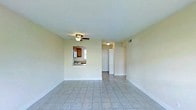This single-floor space features an open and airy living area finished with light-toned ceramic tile flooring throughout, contributing to a clean, cohesive aesthetic. The room is bathed in a soft, neutral palette with cream-colored walls that enhance the brightness of the space. A ceiling fan with a built-in light fixture is centrally mounted, providing both air circulation and ambient lighting.

A defining architectural feature is the pass-through window connecting the living area to the kitchen, allowing natural light from the kitchen's window to filter into the main living space. The kitchen is equipped with warm wood-toned cabinetry and standard appliances, visible through the opening. This pass-through design promotes an open, connected feel between the two areas.

A doorway leads from the living area to additional rooms beyond, suggesting a practical and straightforward floor plan. A wall-mounted vent indicates a central HVAC system. The overall ambiance of the space is understated and functional, with a simple, uncluttered design that offers a solid foundation for personalization and furnishing.