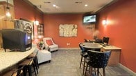 This single-floor communal lounge space makes a strong visual impression with its rich terracotta-red walls, creating a warm and inviting atmosphere throughout. The room is thoughtfully divided into distinct functional zones, catering to both relaxation and productivity. A comfortable seating area features upholstered armchairs with accent pillows, complemented by decorative wall sconces that cast a soft, ambient glow. A large piece of abstract artwork serves as a focal point on the main wall, adding a sophisticated artistic touch alongside additional framed wall décor. For entertainment, a wall-mounted flat-screen television is positioned prominently, making it ideal for casual viewing. On the opposite side, a dedicated workstation area is equipped with computer terminals and high-top bar-style seating with tufted black leather chairs, offering a practical space for business tasks. Neutral-toned carpet tiles cover the floor, providing a subtle contrast to the vibrant walls. Recessed lighting and wall sconces work together to ensure the space is well-lit. Crown molding with a dark trim accent frames the ceiling, adding a refined architectural detail that ties the room's design together. Overall, the space balances comfort, functionality, and bold design.