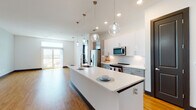 Located at 5126 Peachtree Blvd in Chamblee, Georgia, this single-floor residence offers a thoughtfully designed open-concept layout with modern finishes throughout. The kitchen serves as a centerpiece, featuring an expansive white quartz island with an undermount sink and stylish globe pendant lights suspended overhead. Two-tone cabinetry — upper white and lower grey — pairs seamlessly with a full suite of stainless steel appliances, including a refrigerator, range, and built-in microwave. A clean white tile backsplash completes the polished look. The living area extends naturally from the kitchen and is bathed in natural light from large sliding glass doors that open to an outdoor balcony, offering views of the surrounding area. Warm hardwood-style flooring runs consistently throughout the entire unit, complemented by dark baseboards and dark-framed interior doors that provide a striking contrast to the crisp white walls. The bedrooms are well-proportioned, with large windows, ceiling fans, and ample natural light. One bedroom features decorative wall paneling, adding an architectural detail. The primary bathroom includes a glass-enclosed walk-in shower with tile surround and a walk-in closet, while the secondary bathroom offers a tub-shower combo with built-in open shelving.