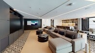 Located at 3200 Paseo Village Way in San Diego, CA, this single-floor amenity lounge offers a refined and contemporary communal space designed for both relaxation and socializing. The space is defined by a striking contrast of dark and light design elements, combining matte black accent walls with crisp white ceilings fitted with recessed and linear LED lighting that creates a warm, layered ambiance throughout.

The main lounge area features generously sized sectional sofas in neutral tones, arranged around bold circular black ottomans, providing ample seating for gatherings. Large floor-to-ceiling windows along one wall flood the space with natural light and offer views of the outdoor surroundings. A dramatic wall mural adds an artistic focal point to this area.

A dedicated media zone anchors one end of the space, complete with a large wall-mounted screen, additional seating, and a long communal table with sleek black chairs — ideal for presentations or casual viewing. Decorative patterned black-and-white tile flooring elegantly transitions between zones, adding visual interest and grounding the overall design.

The wet bar area is a standout feature, boasting rich wood cabinetry, a marble countertop island, mirrored backsplash with under-cabinet lighting, a wine refrigerator, and gold pendant lights that lend a warm, upscale character. A glass-paneled door connects to an adjacent dining or meeting area, enhancing the space's versatility.