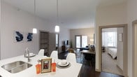 This thoughtfully designed single-floor residence showcases a contemporary open-concept layout that effortlessly connects the dining and living areas, creating a spacious and cohesive environment. The dining area features a sleek white table surrounded by modern white chairs, illuminated by elegant pendant lights that hang from the soaring ceilings, adding a striking visual element to the space. Dark hardwood flooring runs throughout, providing a rich contrast against the light-toned walls and furnishings. The living area is comfortably appointed with a sofa, a patterned area rug, and a media console, while large glass doors at the far end invite an abundance of natural light and offer views to the outside. A built-in shelving unit and additional storage spaces are visible through doorways, highlighting the functional design of the layout. A desk and chair are neatly positioned near the living area, offering a dedicated workspace. Adjacent rooms, accessible through interior doors, include what appears to be a bedroom with soft carpeting, further enhancing the versatility of the floor plan. The overall ambiance is clean, modern, and welcoming, with a neutral palette and carefully chosen furnishings that contribute to a bright and airy atmosphere throughout.