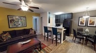 This single-floor residence offers a thoughtfully designed open-concept layout that connects the living and kitchen areas into one cohesive, functional space. The living area features warm hardwood-style flooring, a ceiling fan with light fixture, and a bold yellow accent wall that adds a pop of color and character to the room. A comfortable dark-upholstered sofa, framed wall art, and a rich wood coffee table complete the inviting lounge area.

The kitchen is defined by dark cabinetry that contrasts elegantly with light countertops and a white breakfast bar peninsula, which doubles as a casual dining space with two leather bar stools. Black appliances, including a refrigerator, range, and microwave, lend a sleek, coordinated look to the culinary space. The peninsula provides both additional prep space and a natural division between the kitchen and living areas.

A hallway visible from the main space leads to additional rooms, hinting at a well-organized floor plan. The textured ceiling and neutral wall tones throughout create a balanced backdrop that ties the warm and dark design elements together. The overall ambiance is cozy yet modern, making this a welcoming and practical living environment.