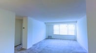 This single-floor space offers a straightforward and functional layout designed to maximize usability. The living area is the centerpiece of the unit, characterized by soft neutral carpeting that extends throughout and clean white walls that create a bright, open atmosphere. A large multi-pane window floods the living space with natural light, offering views to the outdoors while making the room feel airy and welcoming. The smooth, flat ceilings lend a clean, modern aesthetic throughout.

The floor plan, as seen in the dollhouse view, follows an L-shaped configuration that provides natural separation between the living and utility areas. Toward the rear of the unit, a kitchen area is visible with standard cabinetry and fixtures, while a dedicated bathroom space features essential fittings. The transition between areas is seamless, with a consistent use of light tones across walls and surfaces reinforcing the sense of cohesion throughout.

The overall design is minimal and move-in ready, presenting a blank canvas well-suited for personalization. The combination of natural light, open proportions in the main living space, and a practical room arrangement makes this a versatile and functional unit.