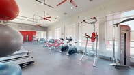 This fitness center offers a spacious, single-floor workout environment designed to accommodate a full range of exercise activities. The interior is defined by a striking red and white color scheme, with bold red accents on equipment, doors, and ceiling fans that create an energizing atmosphere throughout the space.

The facility is outfitted with an extensive lineup of cardio machines, including multiple treadmills, ellipticals, stationary bikes, and stair climbers, arranged in well-organized rows to maximize accessibility. A dedicated strength training area features a full dumbbell rack, adjustable weight benches, cable machines — including a Cybex unit — and a multi-function pull-up and dip station. Stability balls and additional free weights round out the functional fitness offerings.

Large garage-style glass doors on one wall open toward lush greenery outside, flooding the gym floor with natural light and providing refreshing outdoor views. Roller shade windows along the adjacent wall offer additional light control. The polished concrete flooring is durable and easy to maintain, well-suited for a high-traffic fitness environment.

A large wall mirror with a distinctive wood-grain artistic border visually expands the space and aids in exercise form. Recessed lighting and multiple red ceiling fans complete the modern, functional design, ensuring comfort during even the most intense workouts.