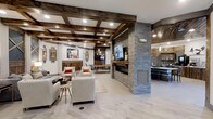 This thoughtfully designed single-floor space seamlessly blends rustic charm with contemporary sophistication. The ceiling immediately draws the eye upward, showcasing a striking coffered design with rich, dark wood beams and recessed lighting that casts a warm, inviting glow throughout the interior.

The main lounge area is anchored by a stacked stone fireplace column, which serves as a dramatic focal point. Mounted above the fireplace is a flat-screen television, making the space well-suited for both relaxation and entertainment. Comfortable neutral-toned armchairs with vibrant orange accent pillows are arranged around a low coffee table, creating an intimate and welcoming seating arrangement.

Adjacent to the lounge, a dining or meeting area is visible, featuring a long wooden table and chairs, ideal for gatherings or collaborative use. The light-toned flooring throughout provides a clean contrast to the darker wood and stone elements, enhancing the sense of openness.

A well-appointed bar area is positioned to one side, complete with pendant lighting, bar seating, and cabinetry, making it a standout feature for social occasions. Rustic wood wall paneling and carefully selected artwork complete the aesthetic, contributing to an atmosphere that is both refined and warmly approachable.