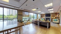 This well-appointed single-floor amenity space is designed to offer a variety of social and recreational experiences within a cohesive, modern interior. Warm hardwood flooring flows throughout, unifying the distinct functional zones. Floor-to-ceiling windows line the perimeter, flooding the space with natural light and framing lush tree canopy and urban views beyond.

A lounge area anchors the space with plush seating in rich gold and earth tones, centered around a fireplace topped by a wall-mounted flat-screen television. Adjacent built-in wood bookshelves add warmth and a library-like character to the setting. Nearby, a dedicated seating nook with high-top bistro tables and striped bar-height chairs takes full advantage of the panoramic window views, creating an inviting spot for casual gatherings.

The space also features a full billiards table, adding a recreational dimension to the layout. A practical kitchenette, outfitted with light wood cabinetry, a sink, and dark countertops, supports events and everyday use. A teal glass-paned door leads to an adjoining area, enhancing connectivity throughout. Recessed lighting and a drop ceiling with inset panels provide consistent, even illumination, contributing to the space's polished and welcoming ambiance.