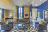 Located at 70 Lakeshore Drive in Hampton, Virginia, this single-story community clubhouse is designed to impress with a bold, contemporary aesthetic. The space is anchored by a striking color palette of deep navy blue, warm gold, and neutral tan, carried consistently throughout via accent walls, drapery, and furnishings.

The main lounge area is expansive and open, boasting soaring ceilings with recessed lighting and eye-catching pendant fixtures. Rich hardwood flooring runs throughout, adding warmth and continuity to the space. Generous windows dressed with floor-to-ceiling navy and gold curtains flood the interior with natural light while offering views to the outdoor surroundings.

Comfortable seating arrangements feature plush blue sofas and accent chairs accented with vibrant yellow throw pillows, creating an inviting and social atmosphere. Colorful patterned area rugs define each seating zone, adding a lively design element underfoot.

A dedicated bar-height counter with stylish wood and metal barstools serves as a social hub, complemented by a wall-mounted flat-screen television set against a mosaic tile backsplash and a built-in coffee station. A separate dining area with dark-finish tables and chairs provides additional gathering space.

The grand entrance features tall arched transom windows and glass-paned double doors, creating a bright and welcoming foyer. Overall, this clubhouse offers a polished, functional, and visually dynamic environment ideal for community gatherings.