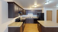 This single-floor unit offers a functional and comfortable layout designed for everyday living. The heart of the space is an open-concept kitchen and living area, where the kitchen stands out with rich, dark-stained cabinetry extending to the upper walls, light-toned countertops, and a full suite of black appliances including a range, refrigerator, and dishwasher. A kitchen island with a wraparound counter provides additional prep space and naturally defines the boundary between the kitchen and the adjacent living area. The kitchen features durable wood-look flooring, while the expansive living area transitions to plush wall-to-wall carpeting, creating a warm and inviting atmosphere. The living space is generously sized, bright with clean white walls, and equipped with baseboard heating. A dedicated entryway with a classic six-panel door welcomes residents into the kitchen area. The bathroom is well-equipped, featuring a dark-stained vanity with storage drawers and cabinets, a light countertop with an integrated sink, a framed mirror with overhead lighting, and easy-care tile-style flooring. The bedroom is comfortably sized with soft carpeting and a baseboard heater, offering a quiet retreat. Overall, the space presents a clean, neutral palette with consistent finishes throughout.