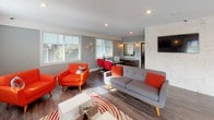 Located at 5700 23rd St E in Fife, Washington, this single-floor commercial space blends modern design with a lively, welcoming atmosphere. The interior is anchored by a bright lounge area furnished with bold orange mid-century modern seating, a sleek grey sofa, and a contemporary coffee table set atop a red-and-white patterned area rug. Recessed lighting and wall sconces complement the abundant natural light streaming through white-trimmed windows with horizontal blinds, highlighting the rich hardwood-style flooring throughout.

A wall-mounted flat-screen television and a clean white credenza define one end of the lounge, while a tiled accent wall adds a refined architectural detail. Adjacent to the lounge, a professional workstation area features a long counter-height desk with red pendant lighting overhead and red adjustable seating, creating a cohesive, energetic color scheme. A windowed pass-through with branded signage connects this workspace to a refreshment station equipped with cabinetry, a coffee maker, and a water dispenser. The open floor plan also accommodates additional functional zones visible in the layout, including what appears to be a fitness or treatment area, making this a versatile, well-appointed space suited for a variety of professional uses.