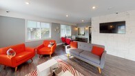 Located at 5700 23rd St E in Fife, Washington, this single-floor commercial space blends modern design with a lively, welcoming atmosphere. The interior is anchored by a bright lounge area furnished with bold orange mid-century modern seating, a sleek grey sofa, and a contemporary coffee table set atop a red-and-white patterned area rug. Recessed lighting and wall sconces complement the abundant natural light streaming through white-trimmed windows with horizontal blinds, highlighting the rich hardwood-style flooring throughout.

A wall-mounted flat-screen television and a clean white credenza define one end of the lounge, while a tiled accent wall adds a refined architectural detail. Adjacent to the lounge, a professional workstation area features a long counter-height desk with red pendant lighting overhead and red adjustable seating, creating a cohesive, energetic color scheme. A windowed pass-through with branded signage connects this workspace to a refreshment station equipped with cabinetry, a coffee maker, and a water dispenser. The open floor plan also accommodates additional functional zones visible in the layout, including what appears to be a fitness or treatment area, making this a versatile, well-appointed space suited for a variety of professional uses.