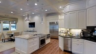 Located at 1411 Parkview Dr in Wilmington, NC, this single-story home showcases refined craftsmanship and thoughtful design throughout. The kitchen stands as a centerpiece of the home, boasting a striking coffered ceiling with recessed lighting and two polished chrome pendant lights that illuminate the space with both style and function. Rich hardwood flooring runs throughout, complementing the bright, white custom cabinetry adorned with classic hardware. The expansive kitchen island is topped with luxurious granite countertops and features an integrated cooktop, wall oven, and warming drawer, making it an ideal hub for cooking and entertaining. A stainless steel built-in refrigerator anchors one wall, while under-cabinet lighting highlights the tile backsplash and additional prep areas along the perimeter. Dark granite countertops on the surrounding cabinetry provide a sophisticated contrast to the white millwork. A built-in television nook adds a modern, convenient touch. The abundance of recessed lighting combined with the bright cabinetry and reflective surfaces creates a warm, welcoming ambiance throughout the kitchen, reflecting the overall quality and attention to detail found in this Wilmington home.