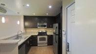 This single-floor space is anchored by a well-appointed kitchen that combines style and functionality. The kitchen features rich, dark espresso cabinetry extending from floor to ceiling, providing generous storage throughout. Light granite countertops create a striking contrast against the dark cabinets, lending the space a clean, contemporary aesthetic. Stainless steel appliances, including a French door refrigerator, electric range, and over-the-range microwave, are seamlessly integrated into the design. Recessed lighting throughout the kitchen ensures bright, even illumination across the workspace. The double-basin sink is positioned along the perimeter, complemented by additional under-counter cabinetry. Warm wood-tone flooring runs through the kitchen, adding a touch of natural warmth to the modern palette. The layout follows an efficient L-shaped configuration, maximizing usable counter and storage space. A glass-paneled entry door is visible at the edge of the space, allowing natural light to filter in. The overall ambiance is modern and practical, with a cohesive dark-and-light color scheme throughout.